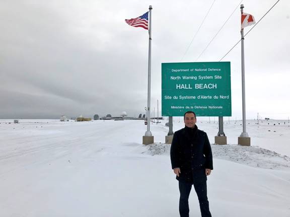 Figure 21 is a photo showing the Chair of the Foreign Affairs Committee standing on the snow outside the site of the North Warning System in Hall Beach. He is standing next to the sign for the site that is flanked by a Canadian flag and an American flag.