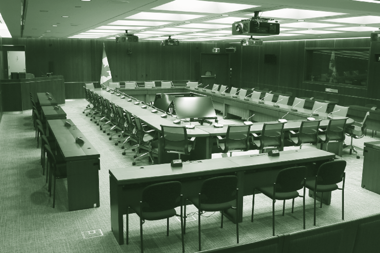 Photograph of a typical committee room, showing several tables placed in a rectangular format with chairs. At the front of the room is a large podium for the Proceedings and Verification Officer (console operator).