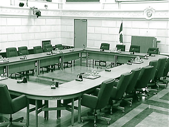 Photograph of a typical committee room, showing several tables placed in a rectangular format, surrounded by chairs. At the front of the room is a large podium for the console operator.